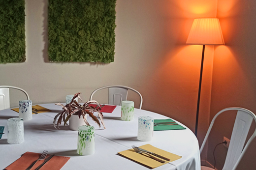 Tavolo di forma tonda, allestita per una cena a base di pizza. Sullo sfondo sono presenti dei quadri rettangolari con del muschio. Nell'angolo è presente una lampada accesa che colora il muro di arancione.